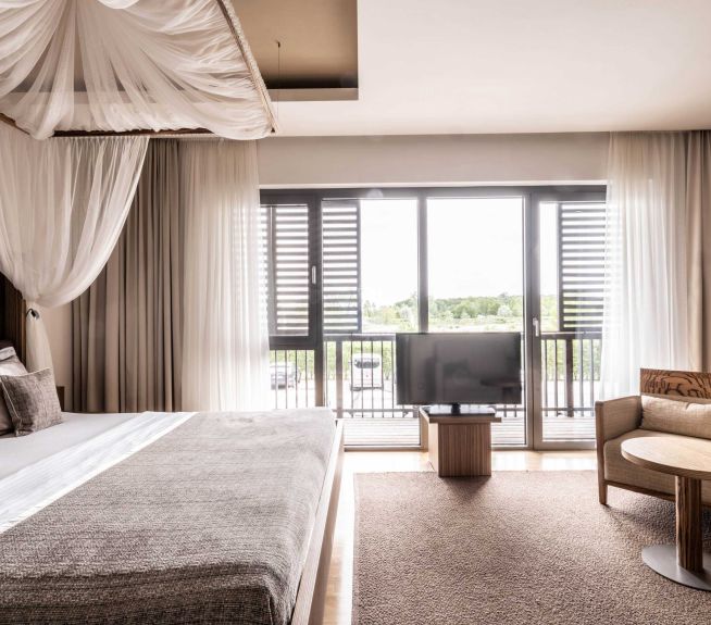 Bright hotel room with four-poster bed, large floor-to-ceiling windows, TV, seating area, and neutral curtains in warm beige tones.