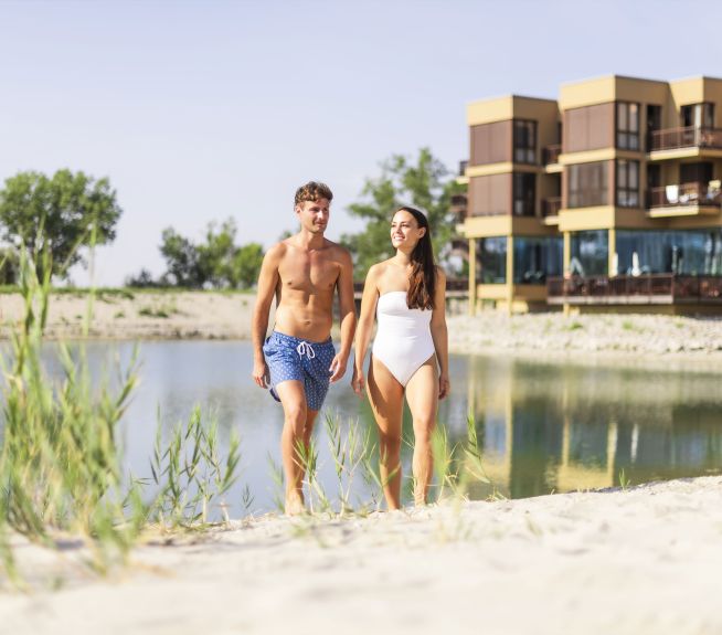 Young couple in swimwear walking along the lodge beach, with St. Martins Therme & Lodge and the lodge's own swimming lake in the background.