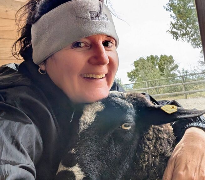 Frau Mag. Schmelzer Elke lächelt und umarmt ein schwarzes Schaf.